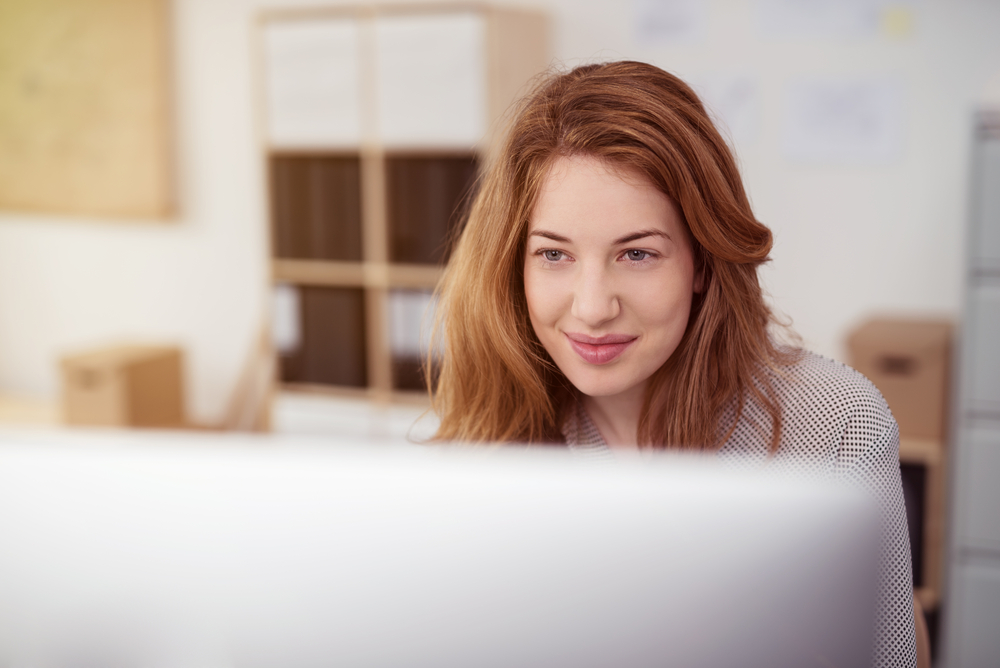 Attractive young woman working on a desktop computer smiling as she leans forwards reading text on the screen, view over the monitor-1 Attractive young woman working on a desktop computer smiling as she leans forwards reading text on the screen, view over the monitor-1